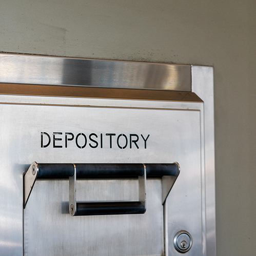 Close-up of a metallic depository box with "DEPOSITORY" printed on it, mounted on a light gray wall, showing a slot for dropping items.