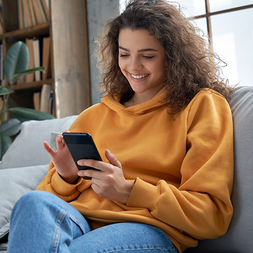 A woman wearing a yellow sweater relaxes on a couch while using her phone.