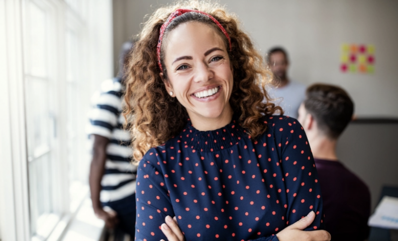 Smiling woman with curly hair wearing a blue polka-dot shirt and pink headband, posing in front of colleagues in an office setting.