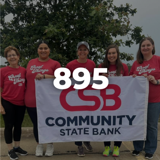 Group of six smiling individuals in red shirts holding a banner for Community State Bank, with the number "895" displayed prominently.
